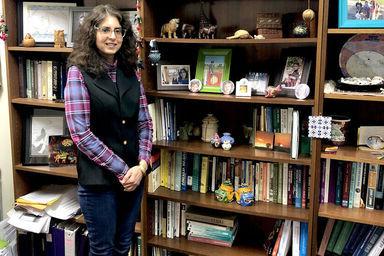 A female professor stands in front of a bookcase in her office filled with books, photos, and different memorabilia 